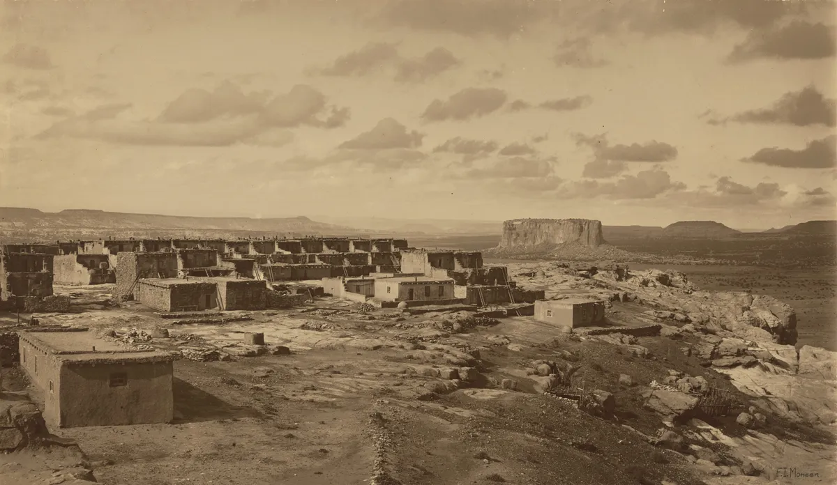 Monochrome photograph of pueblo ruins in a desert mesa landscape. The image has a brownish cast and is rather dreamlike.