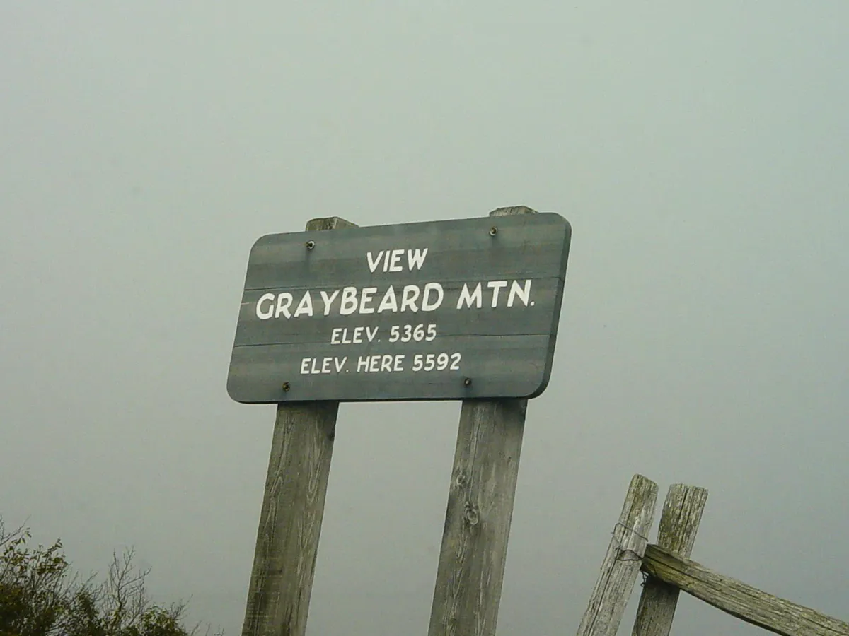 A sign on the Blue Ridge Parkway that reads View Greaybeard Mountain, Elevation 5365 (feet), Elevation Here 5592 (feet). No mountain is visible behind the sign due to dense fog.