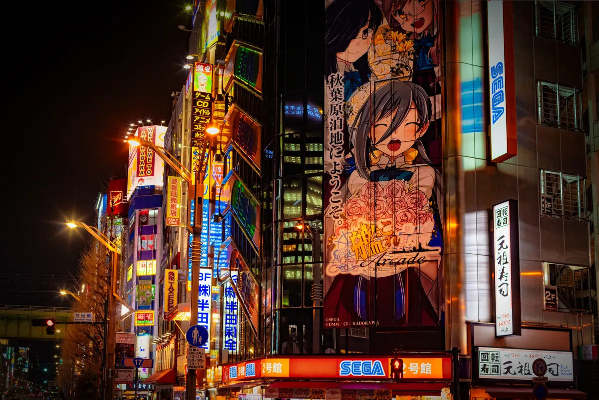 Night scene in Tokyo, skyscrapers; one building has a manga-style illustration of a young woman. Sega logos are prominent.
