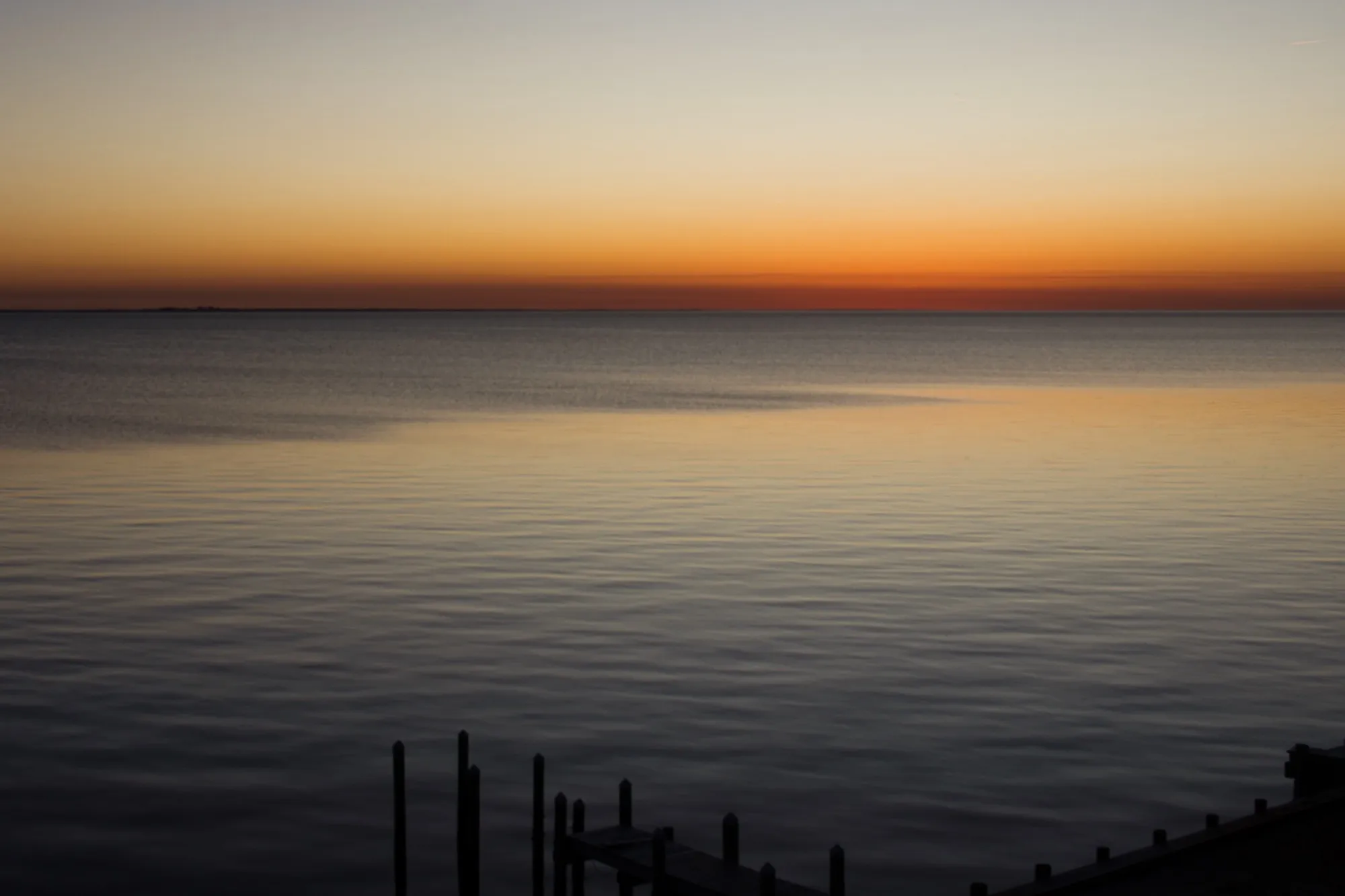 Sunset over a large, mostly still body of water. The sun is already below the horzion, so the water is dark and the sky is fairly dark as well. There are a few wisps of clouds near the horizon that lighten the burt orange of the sky there.