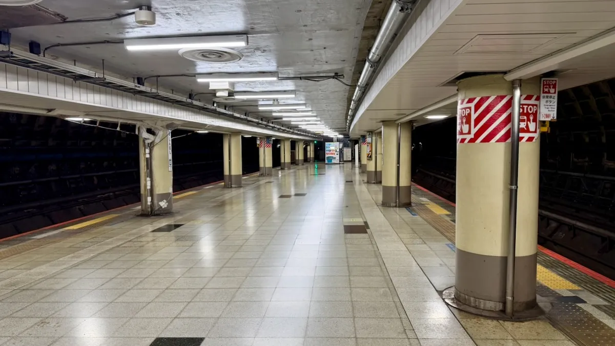 An empty subway platform at Shimashi station in Tokyo; tile floor curves slightly to the right. In the distance you can see a vending machine. Many support pillars have signs saying "stop" on them, which emergency stop buttons.